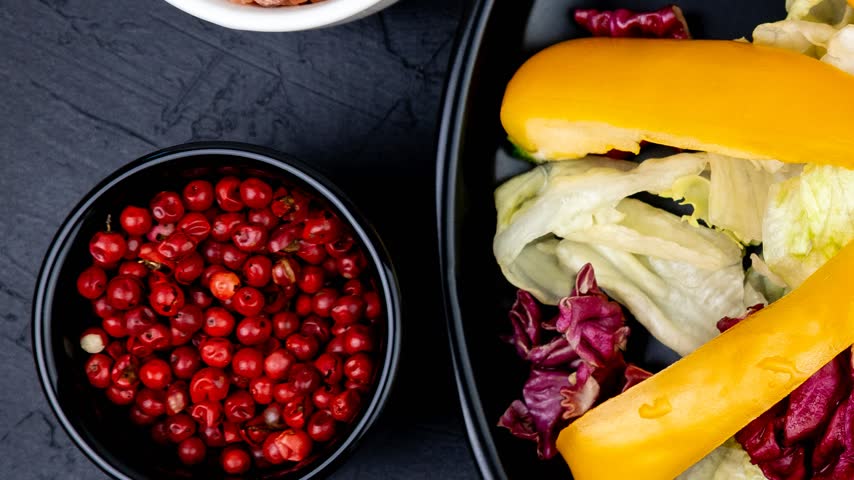 Bowl filled with vibrant red peppercorns placed beside a plate containing mixed lettuce leaves and bright yellow bell pepper slices.