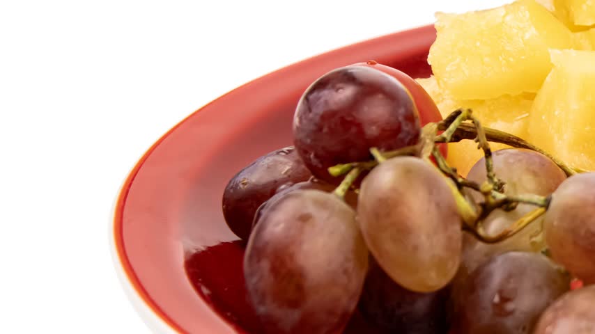 Red grapes with a glossy surface rest on a red plate alongside bright yellow pineapple chunks. The fruits are fresh and vibrant, contrasting nicely with the smooth red plate and the white background.