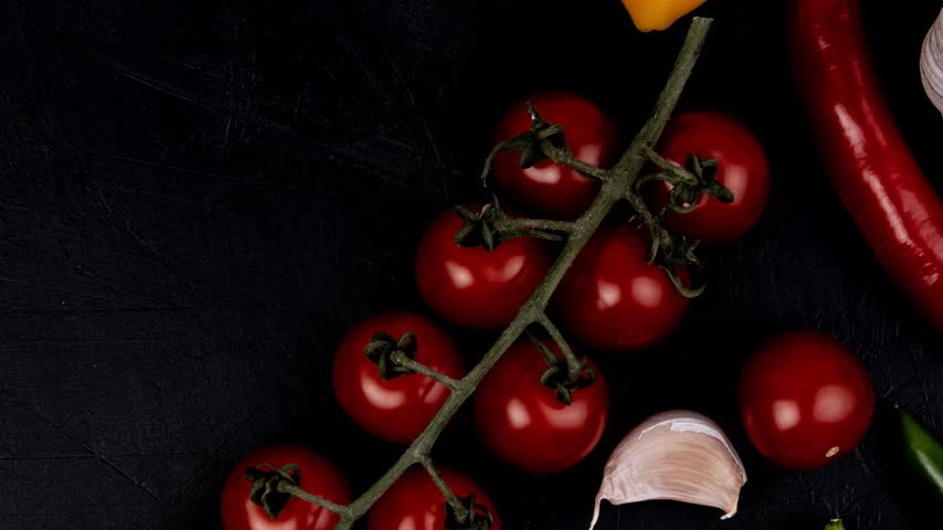 Bright red cherry tomatoes attached to a vine are displayed on a dark textured surface, accompanied by garlic cloves and red chili peppers.