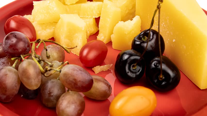 A vibrant cheese plate arrangement featuring chunks of aged cheese, fresh red and black grapes, ripe cherry tomatoes, and glossy black olives on a red plate.