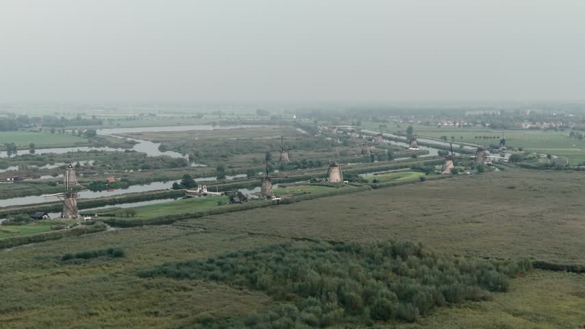 Wide aerial view of flat Dutch countryside with green fields, wetlands, waterways and distant low rise settlements beneath a hazy overcast sky, 4K.