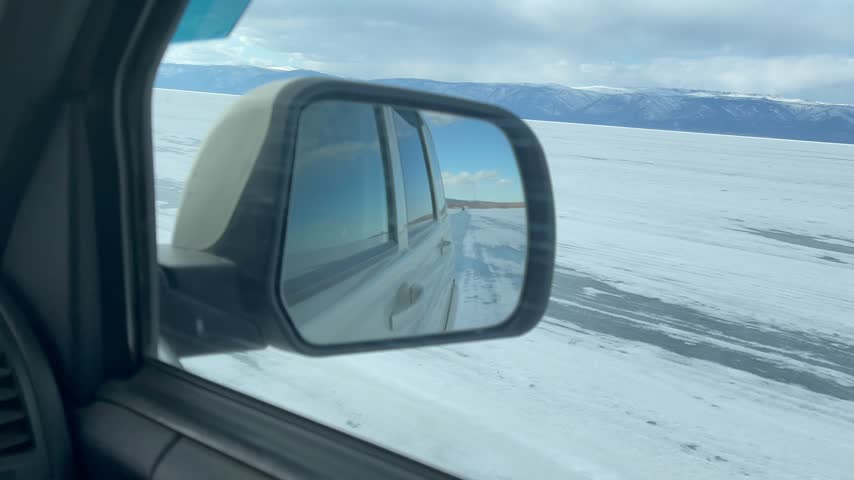 Footage from inside a car driving across the vast frozen ice surface of Lake Baikal in Irkutsk Oblast, Russia, showing the side mirror reflection against snow mountains