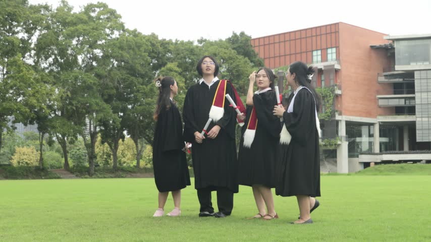 Group male and female university students dressed in graduation gowns and holding their bachelor’s degree diplomas jump high in the air with joy on the campus lawn celebrating their academic achievement.