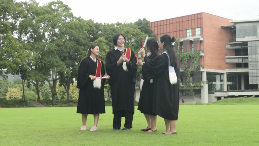 Group male and female university students dressed in graduation gowns and holding their bachelor’s degree diplomas jump high in the air with joy on the campus lawn celebrating their academic achievement.