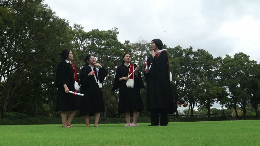 Group male and female university students dressed in graduation gowns and holding their bachelor’s degree diplomas jump high in the air with joy on the campus lawn celebrating their academic achievement.
