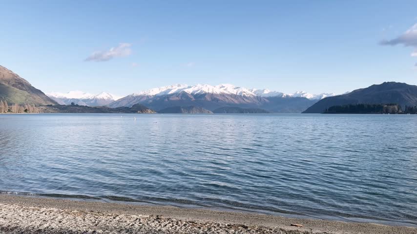 Calm lake water with snowy mountains and clear sky, viewed from a pebbled shore