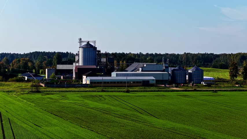 Aerial view of a grain storage facility with metal silos and agricultural buildings surrounded by green fields in Latvia.