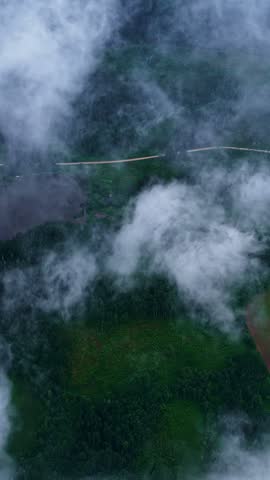 Aerial view of dense forest, a small lake, and a winding road partially obscured by low-hanging clouds in Latvia.