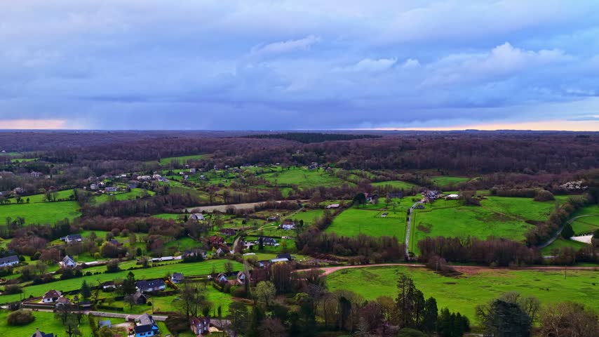 Aerial view of a rural landscape with green fields, scattered houses, and dense forests under a cloudy sky in France.