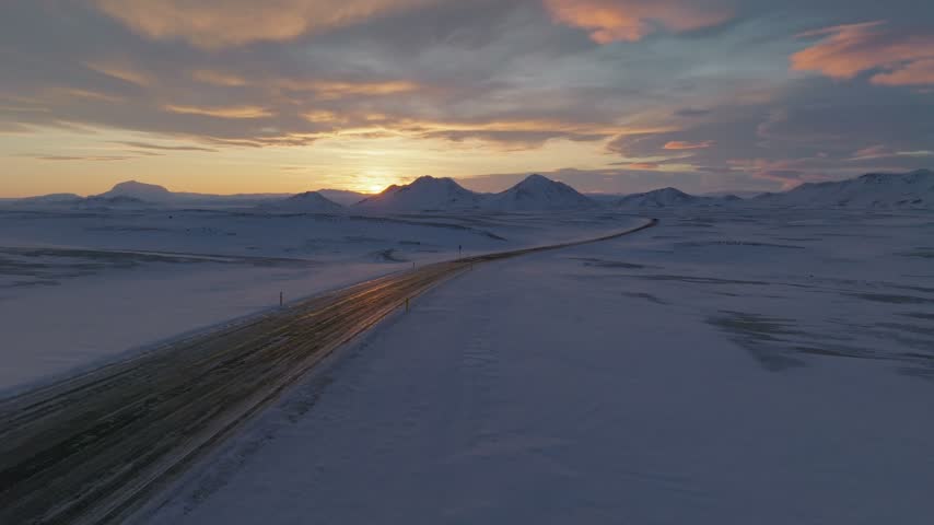 Aerial view of a lonely road stretching through a vast, snow-covered landscape during a golden sunset, with distant mountains under a colorful sky in Iceland.