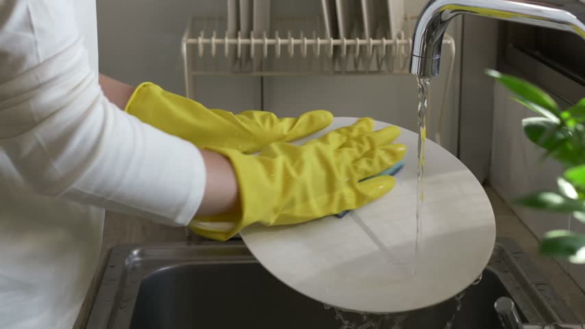 Female hand wearing rubber gloves rotate white plate under running sink water, rinsing its surface. Everyday dishwashing routine promotes kitchen hygiene before clean dishes are placed in drying rack.