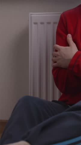 Vertical close-up shot of a cold person wearing a warm red fleece jacket, hugging themselves to stay warm near a radiator heater in an unheated room.