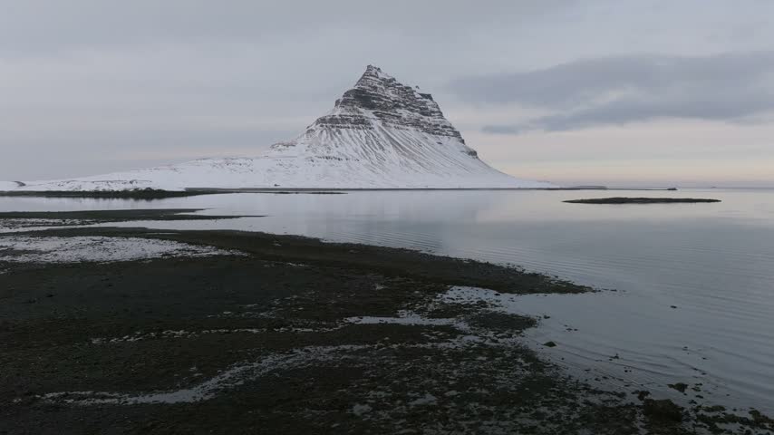 Aerial view of the iconic Kirkjufell mountain covered in snow, rising above the calm coastal waters and dark volcanic shoreline in Iceland.