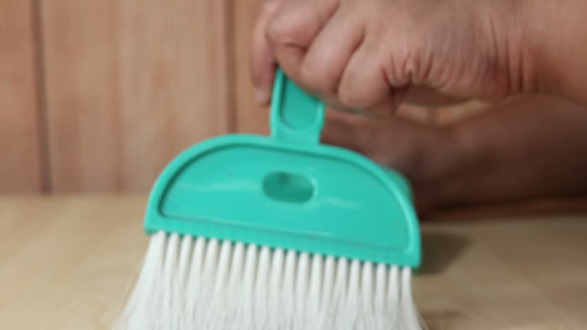 Close up view of hand cleaning wooden desk with compact plastic brush and dustpan set inside bright interior studio room.