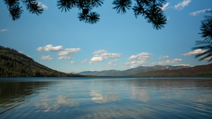 Stanley Idaho wilderness lake timelapse captures gentle ripples on water surface under bright blue sky with moving white clouds and distant forested mountains