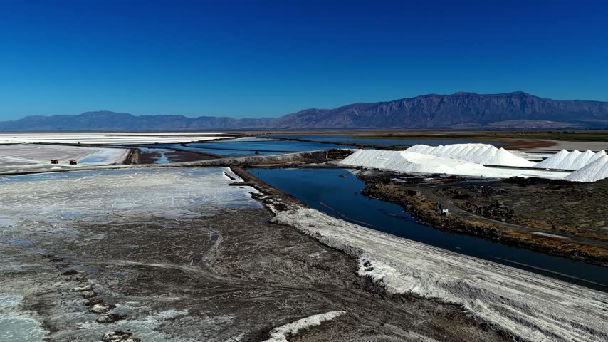 Aerial view of solar salt evaporation ponds along the Great Salt Lake with white salt piles and blue water in Ogden, Utah, United States.