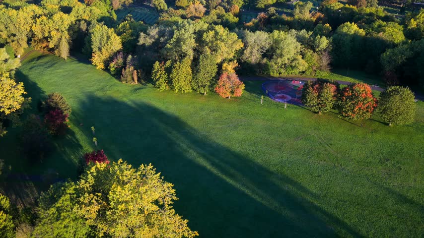 Sunrise aerial views show open grass and trees in Philips Park, East Manchester, United Kingdom, with long shadows across the park.