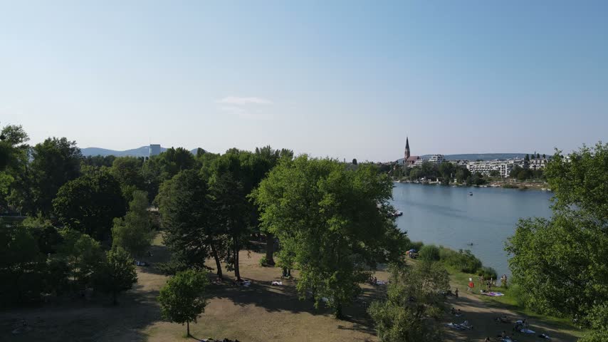 Drone footage of a river flowing through an Austrian town, with green riverside parks, historic buildings and forested hills in the background.
