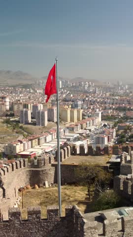 A Turkish flag waves over the inner walls and battlements of Ankara Castle in Ankara, Turkey. The aerial perspective reveals the historic fortifications and surrounding capital city.