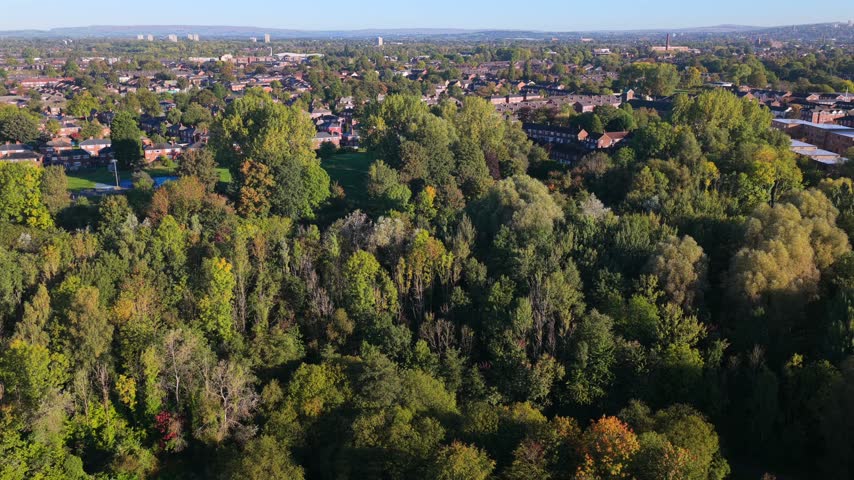 An aerial view looks across Clayton Vale Park and Philips Park in East Manchester, United Kingdom, with trees and homes visible during the morning.