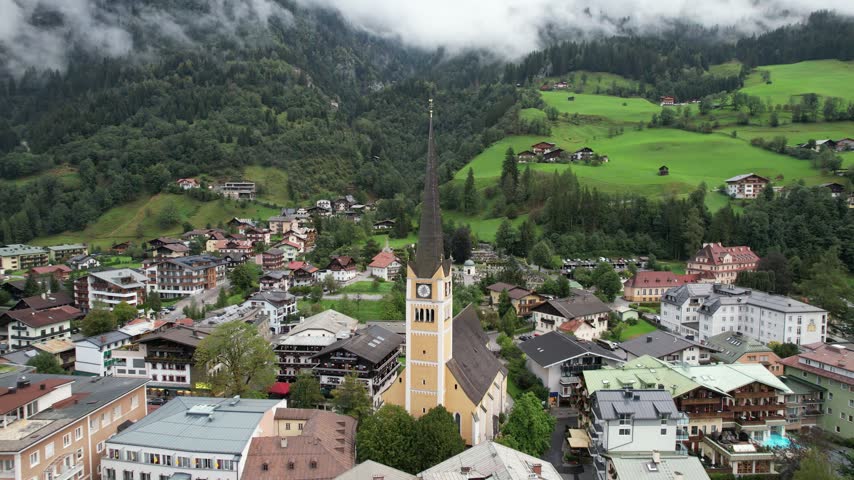 Aerial view of the historic church in Bad Gastein surrounded by traditional alpine architecture, lush green hills, and misty mountains in Bad Gastein, Salzburg, Austria.