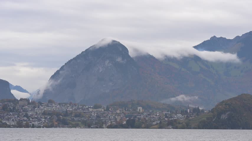 Misty mountains rise behind a lakeside town in Switzerland, representing dramatic alpine weather, peaceful waterfront scenery, European travel, mountain tourism, natural landscape, and scenic.