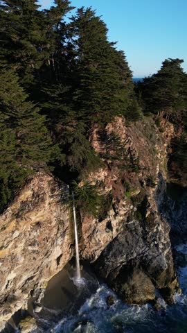 Aerial view of McWay Falls cascading down rugged cliffs onto a sandy beach with turquoise ocean waves in Monterey County, California, United States.