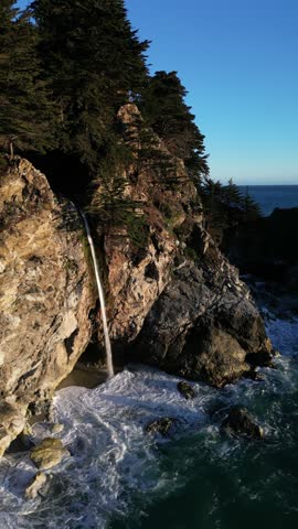 Aerial view of McWay Falls cascading down rugged cliffs onto a sandy beach with turquoise ocean waves in Monterey County, California, United States.