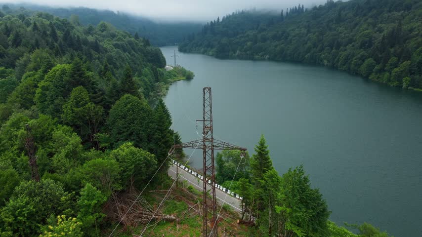Cinematic aerial photography capturing the tranquil mountain Shaori Reservoir in Georgia, tucked away in lush green forest, with road winding along the shore and a visible electric pylon by the water