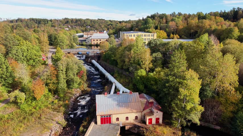 Aerial view of a historic mill building situated by a rushing river surrounded by autumn trees and industrial structures in Fritsla, Vastra Gotaland County, Sweden.