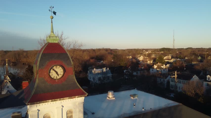 Aerial rising reveal past a historic courthouse clock cupola to small-town rooftops, downtown streets and a water tower in warm winter golden hour light, Denton, Maryland, USA.