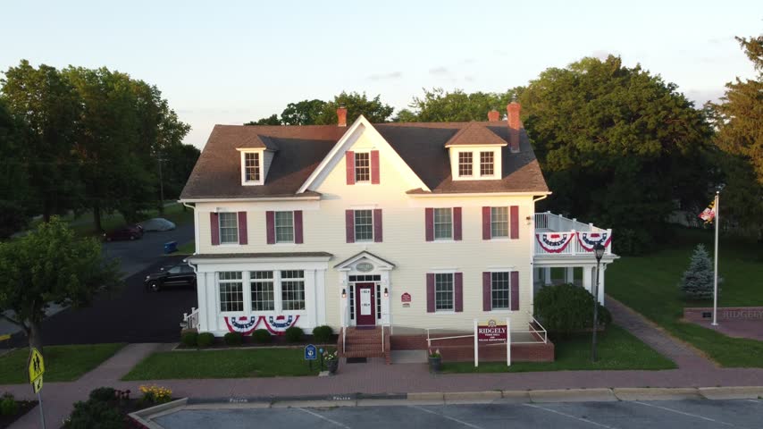 Slow aerial approach toward a white colonial-style town hall decorated with red, white and blue patriotic bunting beside an American flag in golden hour light, Ridgely, Maryland, USA.