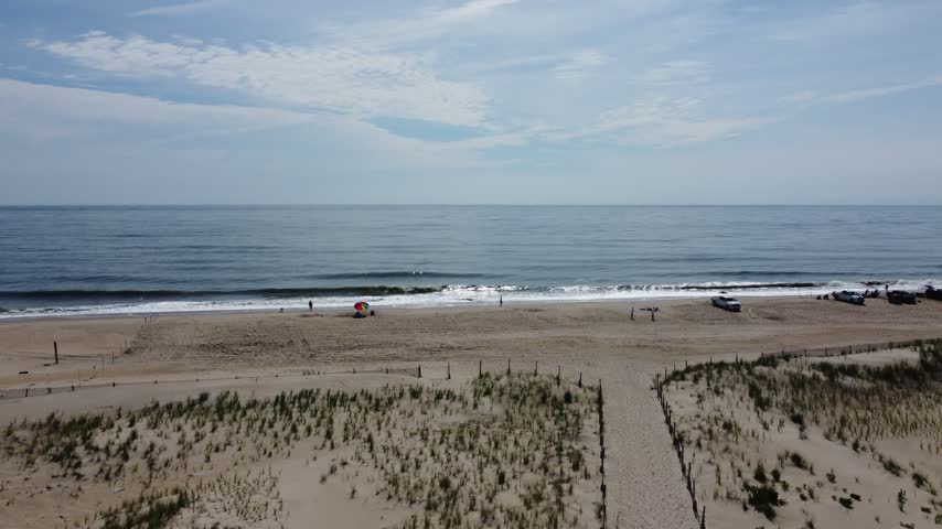 Low hovering drone shot over sand dunes with beach grass and a fenced dune path leading to an ocean beach with distant beachgoers under a summer sky near Lewes, Delaware, USA.