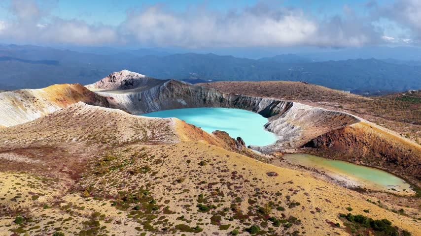 The camera descends from a broad view of layered volcanic ridges toward Yugama Crater Lake, revealing its irregular shoreline and a smaller pond beside the main crater.