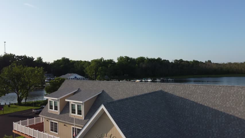Aerial rising reveal over a shingled rooftop opening onto the Choptank River, a marina with docked boats and green marsh vegetation in warm evening light in Denton, Maryland, USA.