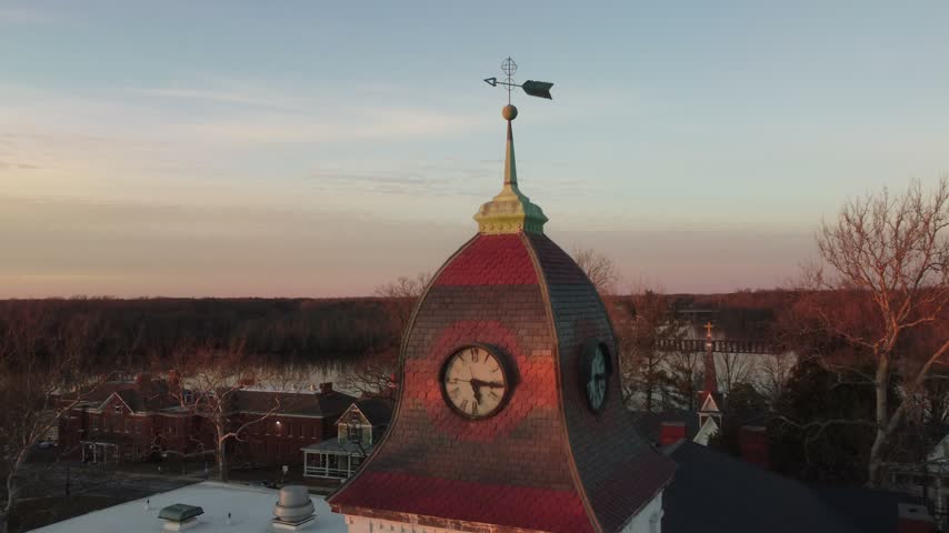 Aerial pullback dronie from a courthouse clock tower cupola and weathervane revealing bare winter trees, the Choptank River and a pastel sunset sky in Denton, Maryland, USA.