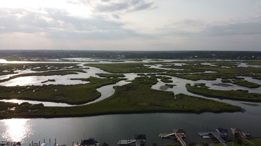 Slow aerial pan across a vast salt marsh laced with winding tidal creeks shimmering with sun glint under a backlit cloudy sky in Garden City Beach, South Carolina, USA.