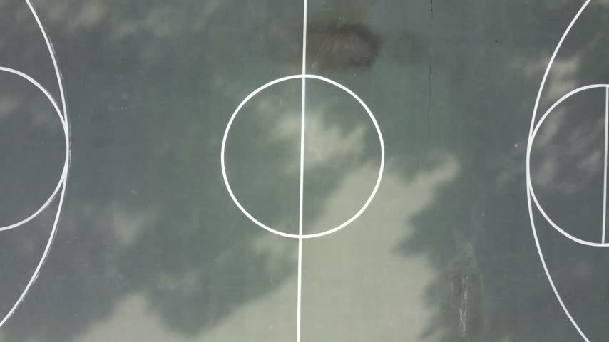 Top-down aerial rising shot from white court lines to a full outdoor basketball court with dappled tree shadows, fencing and grass in a public park in Myrtle Beach, South Carolina, USA.