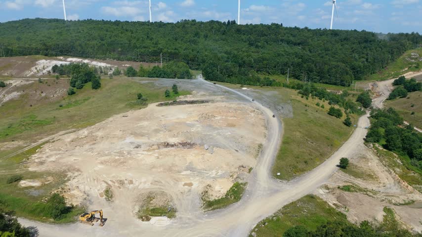 Aerial drone view over a bare hilltop clearing crossed by gravel roads with a line of wind turbines on the forested ridge beyond, Garrett County, Maryland, USA.
