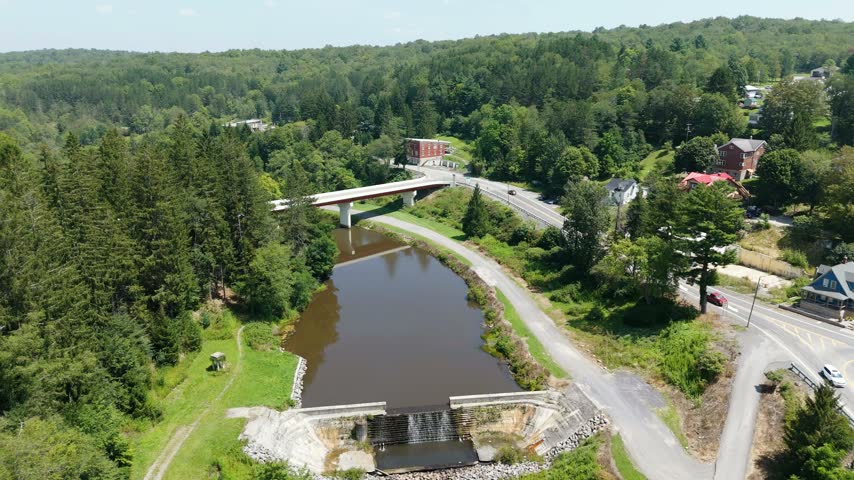 Rising aerial drone reveal over a small dam and pond beside a bridge and road, opening onto forested hills and the town of Thomas, West Virginia, USA.