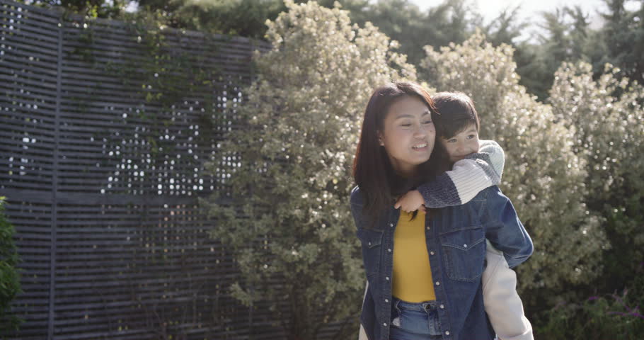 Asian mother in denim carrying son piggyback starting walking in garden by slatted fence bonding. Backyard, child, parent, sunlit, candid, joyful, flowers