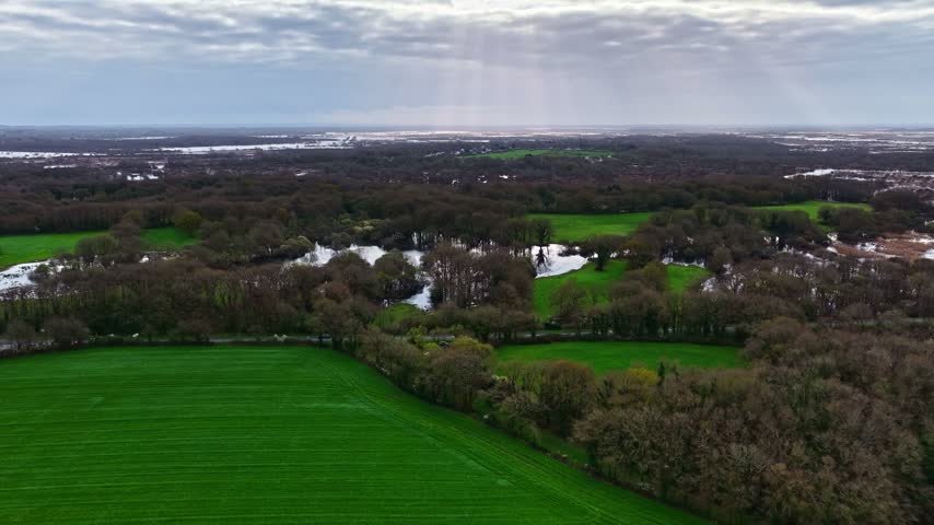 Aerial view of a rural landscape featuring vibrant green fields, patches of forest, and small bodies of water under a cloudy sky in France.