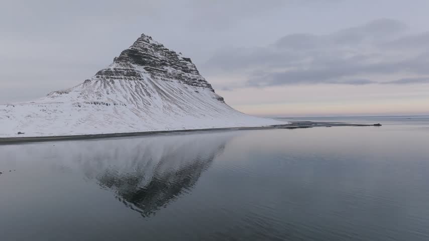 Aerial view of snow-covered Kirkjufell mountain reflected in the calm sea under an overcast sky in Iceland.