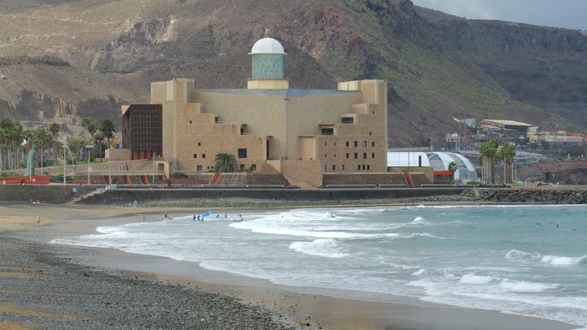 Auditorio Alfredo Kraus concert hall beside Las Canteras beach in Las Palmas de Gran Canaria, with Atlantic waves rolling onto the sand and volcanic hills behind.