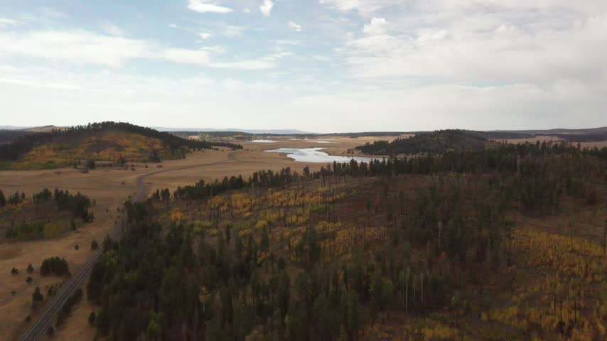 Drone footage captures a scenic landscape with a winding river cutting through open fields and patches of forested hills under a partly cloudy sky. The area features a mix of autumn-colored trees and expansive grasslands, creating a tranquil natural scene.