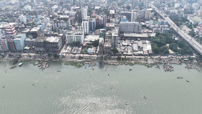 Aerial view of Buriganga River waterfront with numerous small wooden boats moored along the bank, dense urban buildings, and a highway bridge in Dhaka, Dhaka Division, Bangladesh.