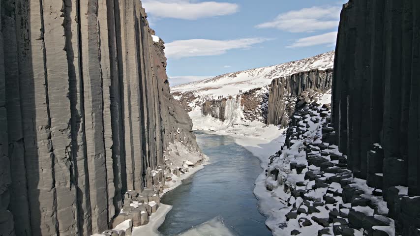 Aerial view of Studlagil Canyon featuring towering basalt columns and a flowing river surrounded by snow-covered landscape in ICELAND.