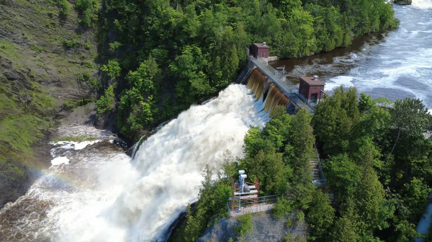 Aerial footage captures a powerful spillway waterfall flowing into a rocky river channel amid dense woodland in Riviere-du-Loup, Quebec, Canada.