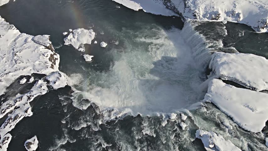 Aerial view of waterfall cascading over snowy cliffs with ice formations and a faint rainbow in ICELAND.