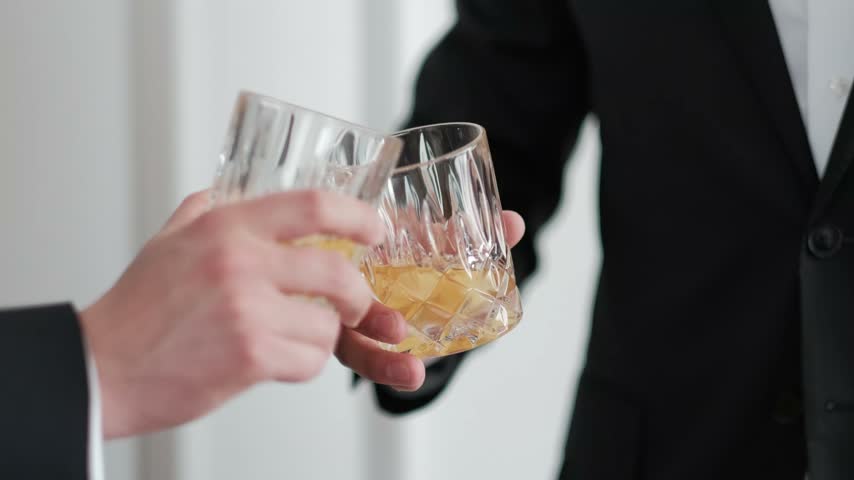 Two men in formal black suits clinking elegant crystal glasses filled with amber whiskey in a toast.